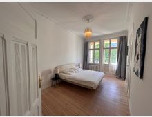 Helles Schlafzimmer mit einem Holzboden, einem Bett mit hellem Holzrahmen und Nachttischen mit Lampen. Große Fenster mit weißen Vorhängen lassen Tageslicht ein und bieten Blick auf draußen; darunter steht ein Heizkörper. Eine Tür mit verzierten Kassetten ist leicht geöffnet.