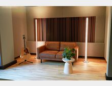 Braun eingerichteter Raum mit einer Leder-Couch, einer Akustikgitarre auf einem Ständer, zwei leuchtenden vertikalen Lampen und einer kleinen Pflanze auf einem weißen Beistelltisch. Der Hintergrund zeigt eine Wand mit schallabsorbierenden Paneelen, der Boden besteht aus hellem Holz.