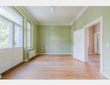 Leerer Wohnraum mit hellgrünen Wänden, hohen weißen Deckenleisten und breiten Dielenholzboden. Links große, hohe Fenster und eine verglaste Tür, rechts eine offene weiße Flügeltür zu einem angrenzenden Raum.