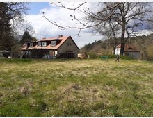 Ein ländliches Grundstück mit einer Wiese im Vordergrund, einem Haus mit braunem Dach und weißen Fenstern in der Mitte, umgeben von Bäumen. Im Hintergrund sind weitere Gebäude und eine Person, die die Szene durchquert.