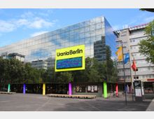 Spiegelglas-Gebäude der Urania Berlin, mit hellgelbem Schild und digitaler Anzeigetafel über dem Eingang. Der Platz davor zeigt Sitzbänke, bunte Säulen und Informationstafeln, flankiert von umliegenden Gebäuden und Bäumen.