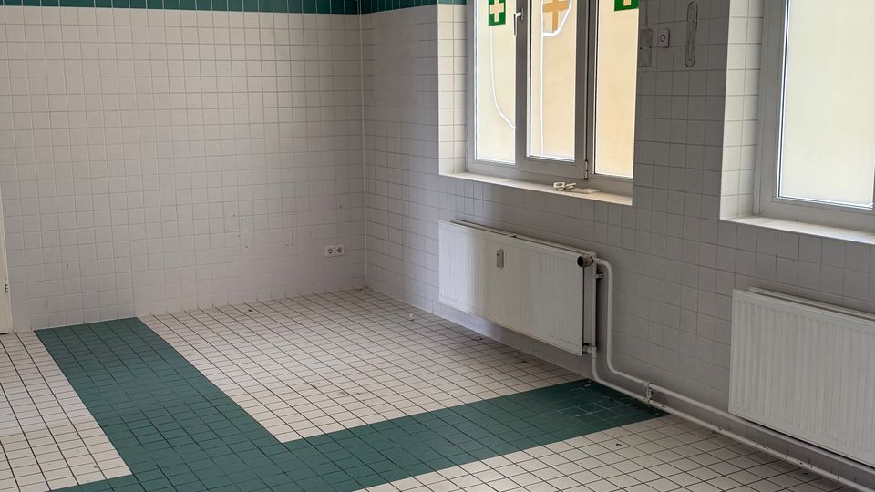 Leerer, gekachelter Raum mit weißen Wandfliesen und einem grünen Fliesenband sowie grün-weiß gemustertem Fliesenboden. Zwei Fenster mit Milchglas und Erste-Hilfe-Kreuz-Symbolen sowie darunterliegende Heizkörper prägen die Seite des Raums.