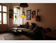 Gemütliches Wohnzimmer mit großer Eckcouch, rundem Couchtisch und Teppich vor hohen Altbaufenstern mit Balkontür. An der Wand hängen Spiegel und Bilder neben einer Gitarre; mehrere Hängepflanzen und eine Stehlampe mit gelbem Schirm prägen die Einrichtung.