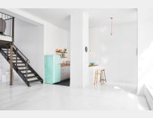 Geräumige, moderne Küche mit einem hellen, minimalistischen Design, weißen Fliesenwänden und einem Retro-Kühlschrank in Pastellgrün; eine schwarze Metalltreppe führt zu einem oberen Bereich, und eine kleine Sitzecke mit Barhockern befindet sich rechts.