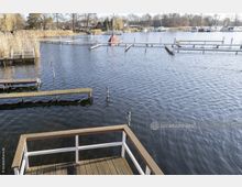 Holzterrassensteg mit Geländer im Vordergrund, Blick auf ein ruhiges Gewässer mit Stegen und vertäuten Booten im Hintergrund; umliegende Vegetation mit Schilf und Bäumen sowie Gebäude am Ufer.