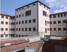 Mehrstöckiges Gebäude mit hellen Fassaden und zahlreichen rechteckigen Fenstern, umgeben von Backsteinmauern mit Stacheldraht; Treppe und Tor befinden sich im Vordergrund. Der Himmel ist teils bewölkt.