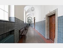 Ein langer Flur mit hellen Gewölbedecken und Wänden, die bis zur halben Höhe mit glasierten, blaugrauen Fliesen verkleidet sind. Auf der linken Seite sind Holzbänke aufgestellt, rechts befindet sich eine Holztür mit Steinrahmen, und am Ende des Flurs ist eine Glastür sichtbar.