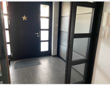 Eingangsbereich mit dunkler Haustür aus Holz und Glaseinsätzen, daneben eine Glastür mit schwarzem Rahmen. Der Boden ist mit grauen Fliesen ausgelegt, an der Wand hängt eine Gegensprechanlage.