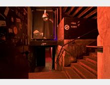 Treppe in einem dunkel beleuchteten Untergrundclub mit Graffiti-beschmierten Wänden und einem aufgehängten Schwarz-Weiß-Porträt. Links eine offene Tür und rechts ein Wegweiser zu verschiedenen Bereichen des Clubs.