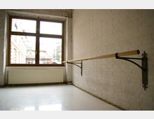 Ein heller Tanzraum mit weiß gestrichenen Ziegelwänden, einem großen Fenster mit Holzrahmen und einer langen Ballettstange an der rechten Wand. Unter dem Fenster befindet sich ein weißer Heizkörper, und der Boden ist aus glänzendem Material.