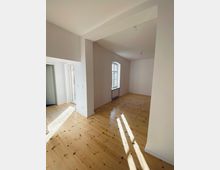 Heller Wohnraum mit hellen Holzdielen, weißen Wänden und großen Fenstern, die viel Tageslicht hereinlassen. Im Hintergrund ist eine Nische mit einem Heizungskörper unter einem Fenster sichtbar.