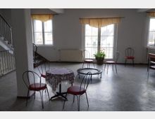 Ein heller Raum mit großen Fenstern, durch die Tageslicht einfällt, ausgestattet mit einfachen Metallstühlen und einem kleinen Tisch mit Spitzentischdecke in der Mitte. Im Hintergrund sind ein Sprossengeländer einer Treppe, ein Heizkörper sowie ein Topf mit Grünpflanzen zu sehen.