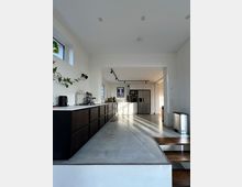 Moderne, offene Küche mit langen dunklen Unterschränken und heller Arbeitsplatte, Betonboden und weißen Wänden. Im Hintergrund stehen hohe Schränke mit Einbaugeräten und ein Edelstahl-Kühlschrank; Deckenstrahler und ein großes seitliches Fenster sorgen für Licht.