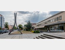 Offener Platz in einer städtischen Umgebung mit modernen Gebäuden und Geschäften, darunter ein flaches Gebäude mit großen Fenstern rechts. Im Vordergrund steht eine abstrakte, hohe Metallskulptur auf einem gepflasterten Boden, umgeben von Grünflächen und parkenden Autos.