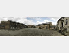 Weitläufiger, gepflasterter Innenhof eines historischen Bauernhofs oder Industrietagwerks, umgeben von mehreren niedrigen, teils verfallenen Gebäuden aus Backstein mit grauen Dächern und weißen Türen. Der Himmel ist bewölkt, und die Szenerie wirkt verlassen.