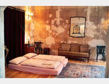 Ein rustikaler Raum mit einer unverputzten Wand voller Abblätterungen, in dem eine einfache Matratze mit Decken und Kissen auf dem Holzboden liegt. Im Hintergrund befinden sich ein antikes Sofa, zwei Stühle, ein Wandspiegel und ein kleiner Teppich, der den Boden teilweise bedeckt.