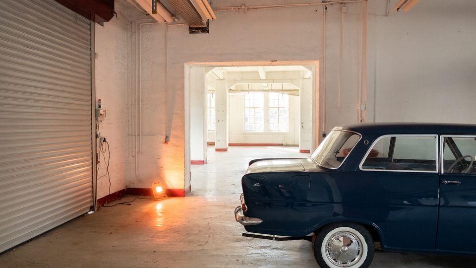 Innenraum einer industriellen Lager- oder Werkstatthalle mit Betonboden, weiß gestrichenen Wänden, sichtbaren Leitungen und einer großen Rolltor-Tür links. Durch eine breite Öffnung führt der Blick in einen hellen Nebenraum mit großen Fenstern; rechts ist ein teilweise sichtbares dunkelblaues Auto geparkt.