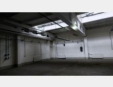 Leerer industrieller Innenraum mit weißen Wänden, freiliegenden Rohren und Kabeln an den Wänden. Die Decke hat große Oberlichter und hängende Leuchtstofflampen; der Boden besteht aus Beton.