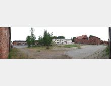 Ein weitläufiger Hof mit Kopfsteinpflaster, umgeben von historischen Backsteingebäuden und einem weißen, mehrstöckigen Gebäude im Zentrum. Im Vordergrund befindet sich eine Wiese mit Bäumen und vereinzelten Bänken; im Hintergrund sind weitere Gebäude und Vegetation sichtbar.