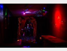 Ein dunkler, stimmungsvoll beleuchteter Raum mit bunter Lichtprojektion an den Wänden und der Decke. Im Vordergrund hängt ein goldener Vorhang, der zu einem Bereich führt, in dem Personen bei schwachem Licht sichtbar sind; rechts ein Tisch mit rotem Stoff und eine rote Lichtquelle.