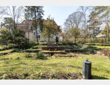 Großer Garten mit Rasenfläche und Blumenbeeten, im Hintergrund ein zweigeschossiges Haus mit Satteldach. Bäume und Büsche umgeben die Szene, rechts steht ein Pavillon aus Metall.