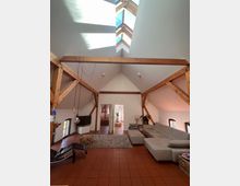 Wohnzimmer in einem Dachgeschoss mit hohen, weißen Wänden und sichtbaren Holzbalken. Der Raum verfügt über ein großes Oberlichtfenster, rote Fliesen, eine graue Eckcouch, eine Liege, und moderne Dekorationselemente.