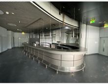 Innenansicht eines leeren modernen Raumes mit einer geschwungenen Metall-Bar in der Mitte, hängenden zylinderförmigen Lampen und einer dunklen Decke mit perforiertem Design. Im Hintergrund sind Türen, ein Notausgangsschild und silberne Wandverkleidungen erkennbar.