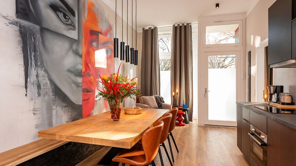 Modernes Wohnküchen- und Esszimmer mit großem Holztisch und Lederstühlen, schwarzer Einbauküche und hängenden Zylinder-Pendelleuchten. An der linken Wand befindet sich ein großformatiges Wandbild, am Ende des Raums eine hohe Glas-/Terrassentür mit Vorhängen und viel Tageslicht.