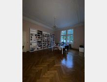 Großes Arbeitszimmer mit hellem Holzparkett in Fischgrätenmuster, einer Bücherwand aus weißen Regalen, einem großen Schreibtisch mit Bürostühlen und einem Kronleuchter an der Decke. Große Fenster lassen Tageslicht herein und bieten Blick auf eine grüne Außenszene.