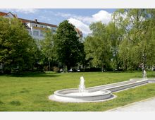 Grüne Parkanlage mit einer großen Rasenfläche, schattenspendenden Bäumen und einem niedrigen, weißen Brunnen mit Wasserfontänen im Vordergrund. Im Hintergrund sind mehrstöckige Wohngebäude mit roten Dächern zu sehen.