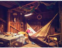 Ein rustikaler Innenraum aus Holz mit Holzbalken und Wanddekorationen wie einem Paddel und einem Rettungsring. Im Vordergrund steht ein gedeckter Tisch mit einer karierten Tischdecke sowie geflochtene Korbstühle, während eine Hängematte unter einer warmen Lichtquelle hängt.