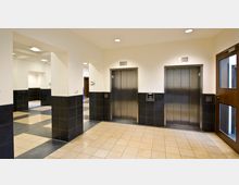 Flur mit zwei geschlossenen silberfarbenen Aufzugtüren, umgeben von schwarz gefliesten Wänden im unteren Bereich und weißen Wänden oben. Der Boden ist beige-schwarz gefliest, und rechts befindet sich eine Glastür mit Holzrahmen.