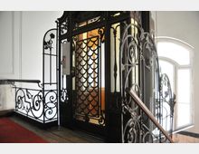 Historischer Aufzug mit kunstvoll gestalteten Schmiedeeisen-Elementen und dekorativen Gittern, umgeben von einem hellen Treppenhaus mit weißen Wänden und einem roten Teppich auf dem Boden. Im Hintergrund sind Fenster mit Tageslichteinfall zu sehen.