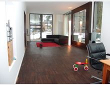 Moderner Wohnraum mit Holzfußboden, einer großen Glasfront mit Blick auf einen verschneiten Garten, einem schwarzen Sofa auf einem roten Teppich und einzelnen Möbelstücken wie einem Schreibtischstuhl und einem Kinderfahrzeug. Links im Vordergrund ist ein Regal mit einer Stereoanlage sichtbar.