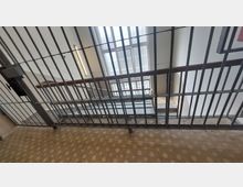 Treppenhaus mit Metallgitter vor den Treppen, welches den Zugang sperrt. Im Hintergrund ein großes Fenster mit Milchglas, das Tageslicht hereinlässt, und Wände in heller Farbe.