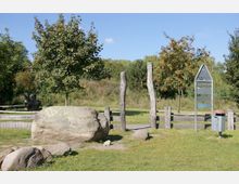 Naturpark mit Grasflächen, vereinzelten Bäumen und einem großen Stein im Vordergrund; im Hintergrund eine kleine Holzkonstruktion als Tor sowie eine Pfosten-Barriere. Ein Mülleimer und ein Infoschild aus Metall befinden sich rechts im Bild.