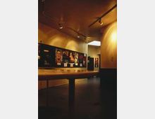 Ein moderner Innenraum eines Museums oder einer Ausstellung mit einer beleuchteten Wand, an der Bilder und Beschreibungen angebracht sind. Der Raum hat eine Decke mit Spotbeleuchtung und helle, minimalistische Gestaltung.