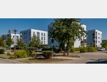 Moderne Wohnanlage mit mehreren weißen Mehrfamilienhäusern, die vier Stockwerke hoch sind und Balkone besitzen. Davor befinden sich Grünflächen mit Bäumen und Sträuchern, vereinzelte geparkte Autos sowie gepflasterte Gehwege und Straßen.