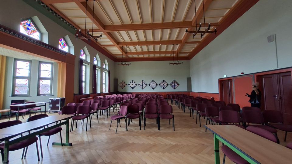 Großer Veranstaltungssaal mit Parkettboden und vielen in Reihen aufgestellten bordeauxfarbenen Stühlen, seitlich ergänzt durch lange Tische. Hohe Fenster mit farbigen Glasfeldern, eine holzverkleidete Kassettendecke mit hängenden Leuchtern und eine Rückwand mit geometrischen Wandtafeln prägen den Raum.