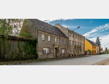 Straßenszene mit älteren, zweigeschossigen Wohnhäusern, teils verputzt, teils mit verwitterten Fassaden; links ein bewachsenes Holzgebäude, rechts farbige Gebäude in Gelb und Rot. Der gepflasterte Bürgersteig und die Straße werden von vereinzelten Straßenlaternen gesäumt.