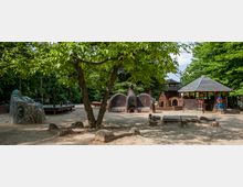 Ein Spielplatz mit sandigem Untergrund, umgeben von Bäumen, enthält fantasievolle, künstlerisch gestaltete Spielflächen, darunter bemalte Keramikelemente, ein Holzdachpavillon und eine Sitzbank aus Mosaik. Im Hintergrund sind kleine, künstlerische Hütten und Figuren integriert.