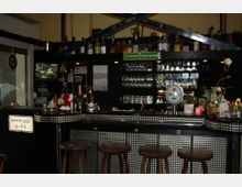 Ein kleiner, gemütlicher Bartresen mit metallisch gemusterter Verkleidung, dekoriert mit Flaschen, Gläsern und Accessoires. Im Hintergrund sind Regale mit mehreren Reihen von Gläsern sowie verschiedene Spirituosen und Dekorationen sichtbar.