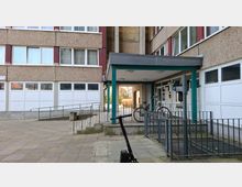 Eingangsbereich eines mehrstöckigen Wohnblocks mit rauer Fassadenstruktur, großen Fensterreihen und einer überdachten Vorfahrt mit grünen Stützen. Davor liegen gepflasterte Wege mit Rampe und Geländern sowie ein Fahrradabstellbereich neben den Haustürbriefkästen.