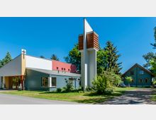 Modernes Kirchengebäude mit flachem Dach und farbiger Fassade, daneben ein freistehender, schmaler Glockenturm mit Holzelementen. Im Hintergrund sind Bäume und ein traditionelles Haus mit Holzfassade zu sehen.