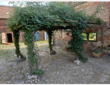 Holzpergola mit dichtem Efeubewuchs, umgeben von einem Hof mit unebenem Steinboden. Im Hintergrund sind ruhige, historische Backsteingebäude mit offenen Türen und kleinen Fenstern zu sehen.