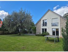 Ein Einfamilienhaus mit heller Fassade und Teilen aus Klinkerstein, zwei Balkonen und großen Fenstern. Im Vordergrund ein gepflegter Garten mit einem großen Baum und einer Tischtennisplatte, umgeben von Büschen und Rasen.