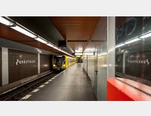 U-Bahn-Station "Pankstraße" mit einem ankommenden gelben Zug auf der linken Seite. Die Wände sind mit braunen Fliesen verkleidet, das Deckenpaneel ist aus Holz, und auf der rechten Seite befinden sich reflektierende metallene Oberflächen.
