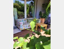 Terrassenszene mit einem bunten Sofa mit floralem Muster, einem kleinen Holztisch daneben und Topfpflanzen, die teilweise eine graue Wand bedecken. Im Hintergrund Glas-Schiebetüren, die zu einem Innenraum führen, und ein Blick auf Bäume im Garten.