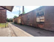 Industriegelände mit alten, roten Backsteingebäuden. Die Fassade zeigt große, teils verrostete Fenster und Türen, daneben verläuft ein Gleis auf gepflastertem Boden; im Hintergrund sind eine Schornsteinspitze und Bäume sichtbar.