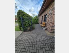 Gepflasterter Eingangsweg neben einem Backsteinhaus mit Dachüberstand und Fenster, an der Wand steht eine Holzbank mit Pflanzkübel. Links befindet sich ein schwarzes Metalltor mit Hecken und Sträuchern entlang der Grundstücksgrenze.